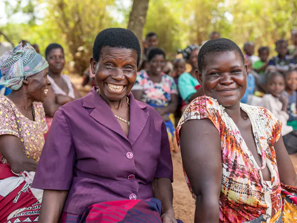 Two smiling Malawian sit in amongst their community under some trees